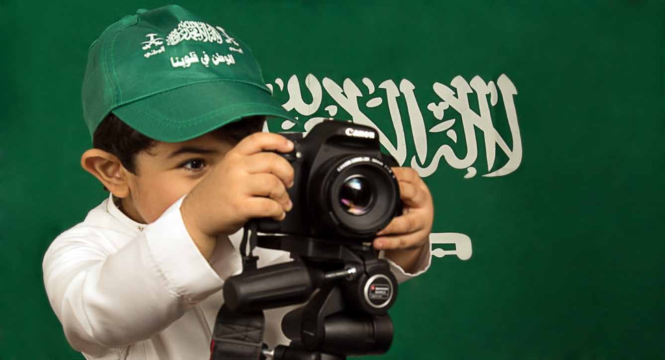 مصور شاب يحمل كاميرته أمام علم المملكة